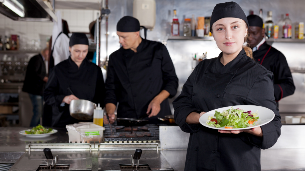 Equipa de cozinha profissional em ambiente de restauração, com vários cozinheiros a trabalhar ao fundo e uma cozinheira em primeiro plano a segurar um prato de salada, junto a equipamentos industriais em inox.