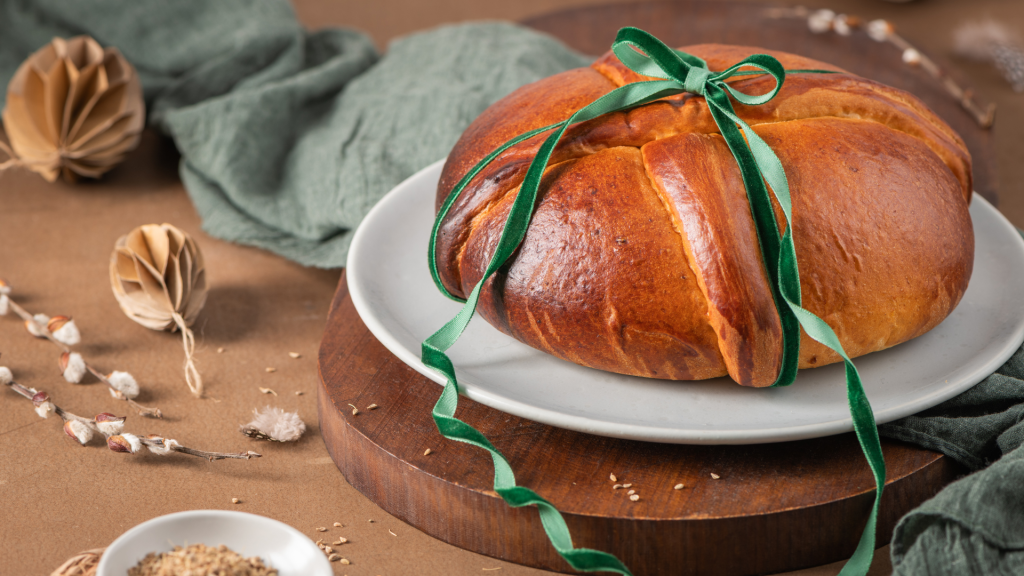 Folar da Páscoa dourado e entrançado, decorado com uma fita verde, servido num prato branco sobre base de madeira, com elementos rústicos em fundo.