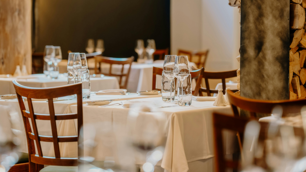 Sala de restaurante elegante e acolhedora, com mesas postas de toalha branca, copos de vinho e cadeiras de madeira, num ambiente intimista e bem iluminado.