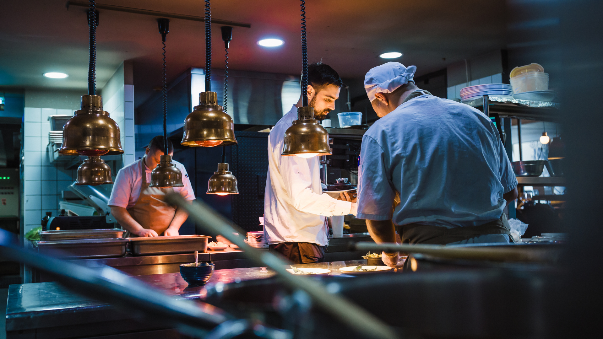 Cozinha profissional de restaurante em plena operação, com chefs a trabalhar nos bastidores, evidenciando fluxos, equipamento técnico e eficiência no setor HORECA.