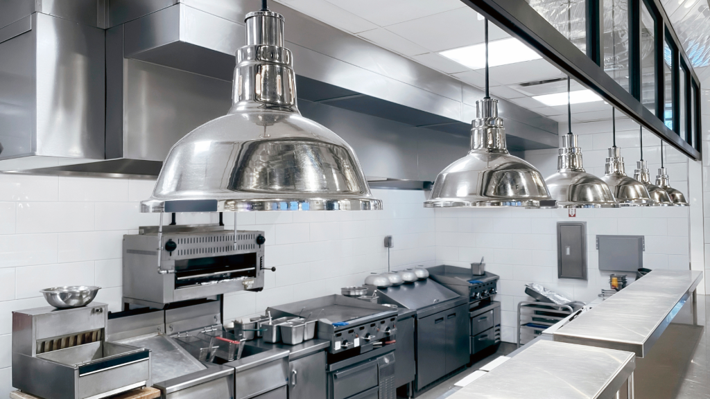 Cozinha profissional em aço inoxidável, equipada com zonas de confeção, exaustão, bancadas técnicas e iluminação suspensa, num ambiente limpo e organizado.