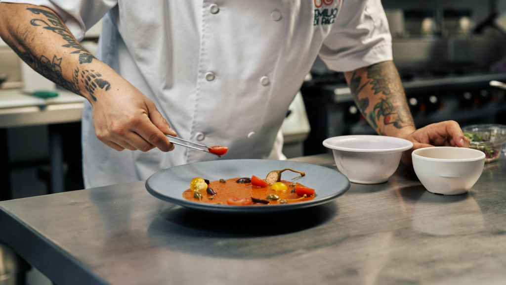 Chef emprata cuidadosamente legumes numa cozinha profissional, utilizando uma pinça para finalizar a apresentação do prato.