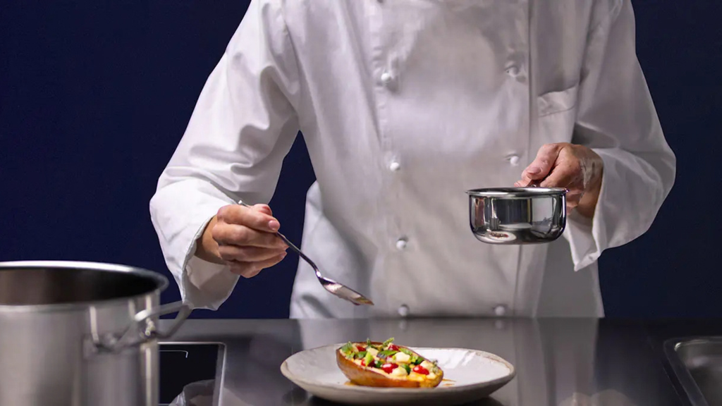 Chef com jaleca branca finaliza um prato com legumes numa cozinha profissional, segurando uma pequena panela e uma colher.