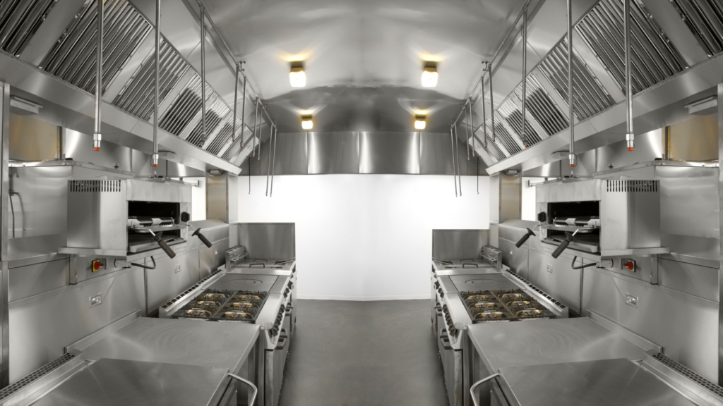 Cozinha profissional com bancadas e equipamentos de confeção em inox, dispostos em duas linhas paralelas, com hottes de extração e corredor central livre.