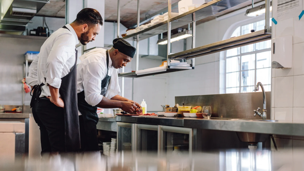 Dois cozinheiros numa cozinha profissional, um a preparar alimentos e outro a observar, junto a uma bancada em inox com arrumação inferior e prateleiras suspensas.